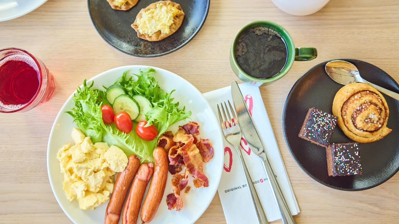 Aamiaispöytä, jossa lautasella munakokkelia, makkaraa, pekonia, salaattia ja tomaatteja. Vieressä kahvi, pulla, browniet ja punainen juoma.