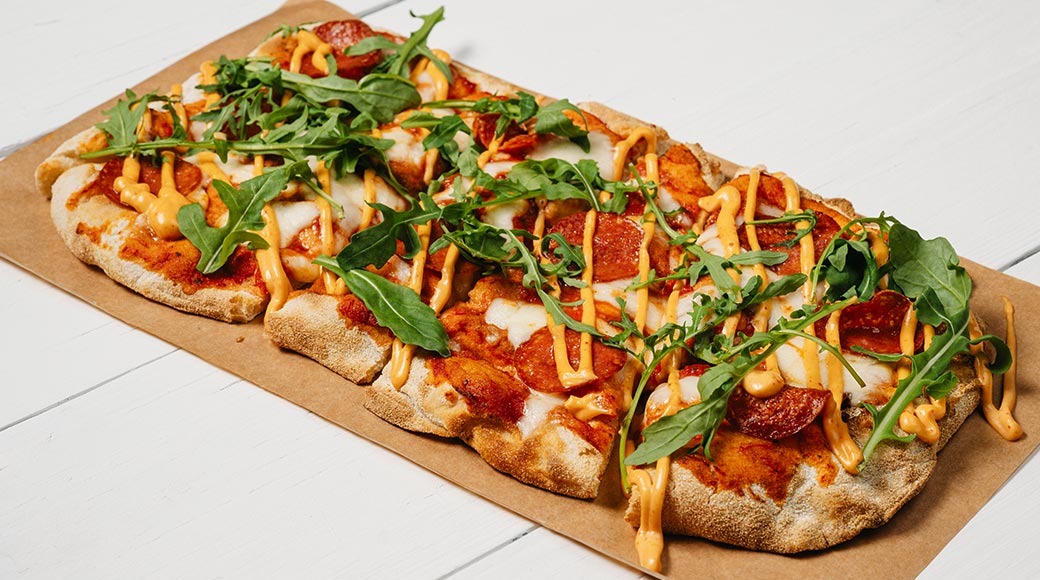 Ristkülikukujuline pepperoni pitsa, mille peal on sulatatud juust, rukola ja oranž kaste. Pitsa on pruunil paberil valgel puitlaual.