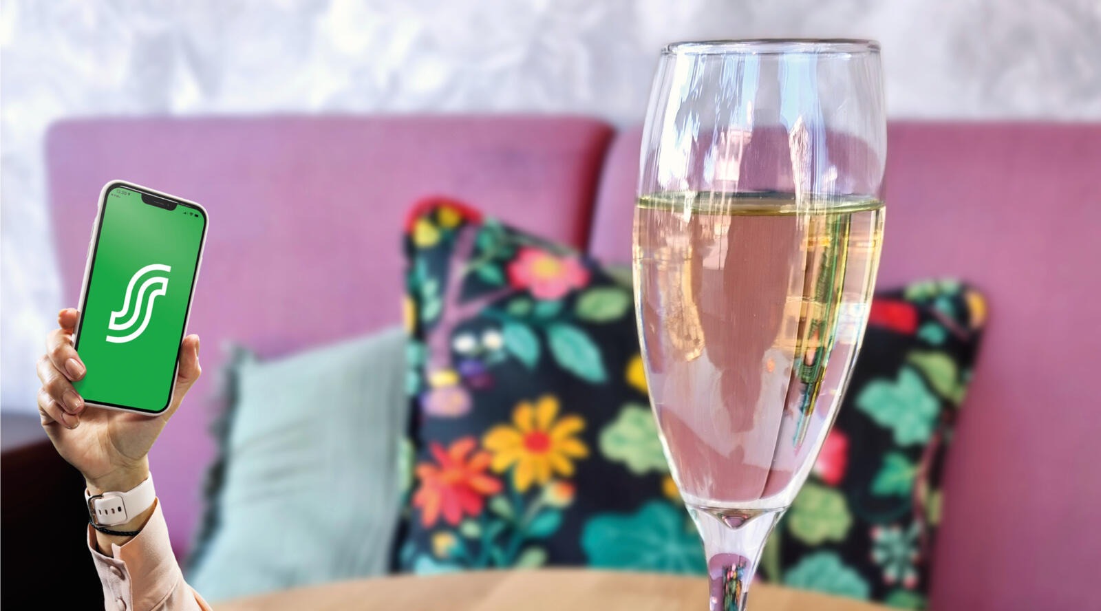 Hand holding a phone in front of a pink sofa, champagne glass on the table.