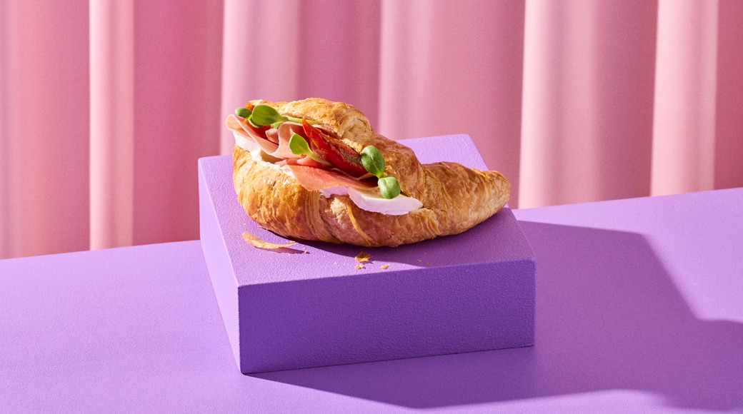 Voisarvi, jonka välissä on kinkkua, juustoa, tomaattia ja versoja, violetin korokkeen päällä vaaleanpunaista verhoa vasten.