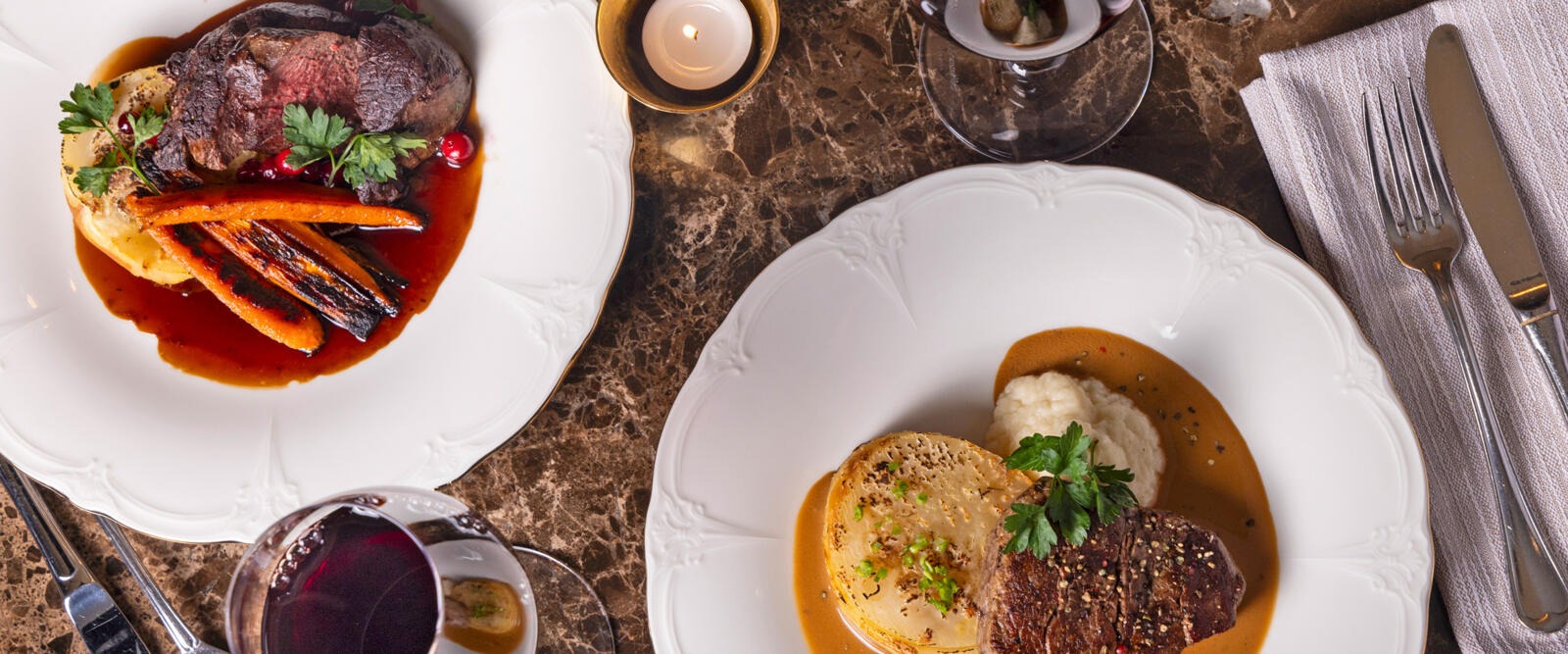Två eleganta tallrikar med mat på ett marmorbord, i ljuset av ett stearinljus. På tallrikarna finns biffar, grönsaker och såser.