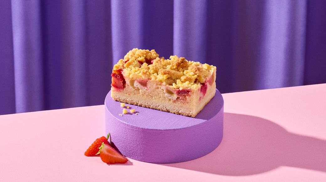 Mansikka-raparperipiirakan pala murupinnalla violetilla korokkeella vaaleanpunaisella pöydällä. Vieressä mansikan puolikkaita.