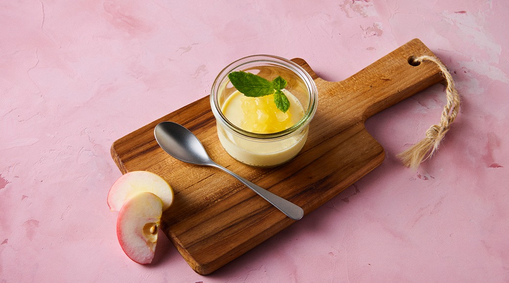 Creamy dessert in a glass jar topped with apple compote and mint. The jar sits on a wooden cutting board next to a spoon. Two apple slices lie nearby on a pink surface.