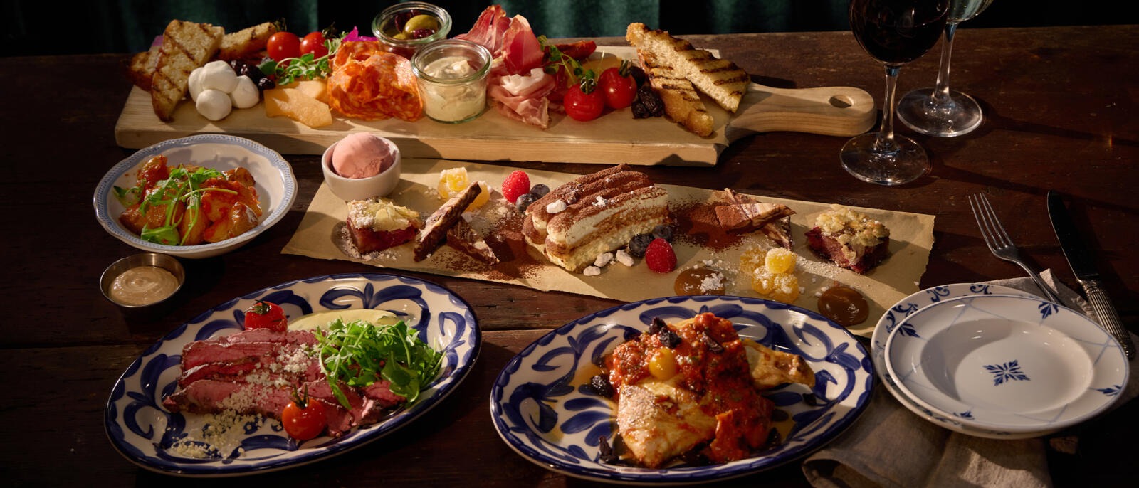 Runsas ateriakokonaisuus, jossa leikkuulauta täynnä juustoja ja leikkeleitä, jälkiruokavalikoima, seared lihaa, kalaa tomaattikastikkeessa ja viinilaseja puupöydällä.