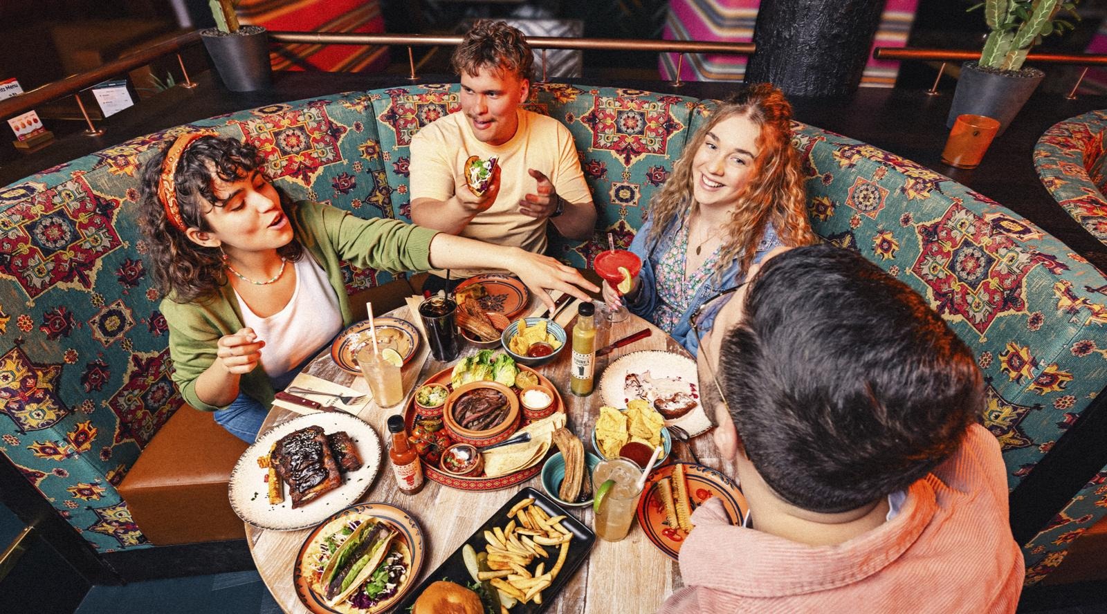 Four people sitting in a colorful patterned booth at a restaurant, table filled with various food plates and drinks.