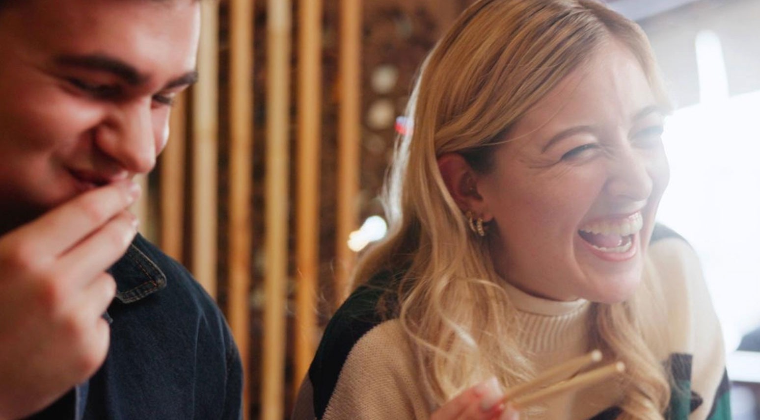 Två personer skrattar tillsammans i ett varmt utrymme med träpaneler i en ljus miljö.