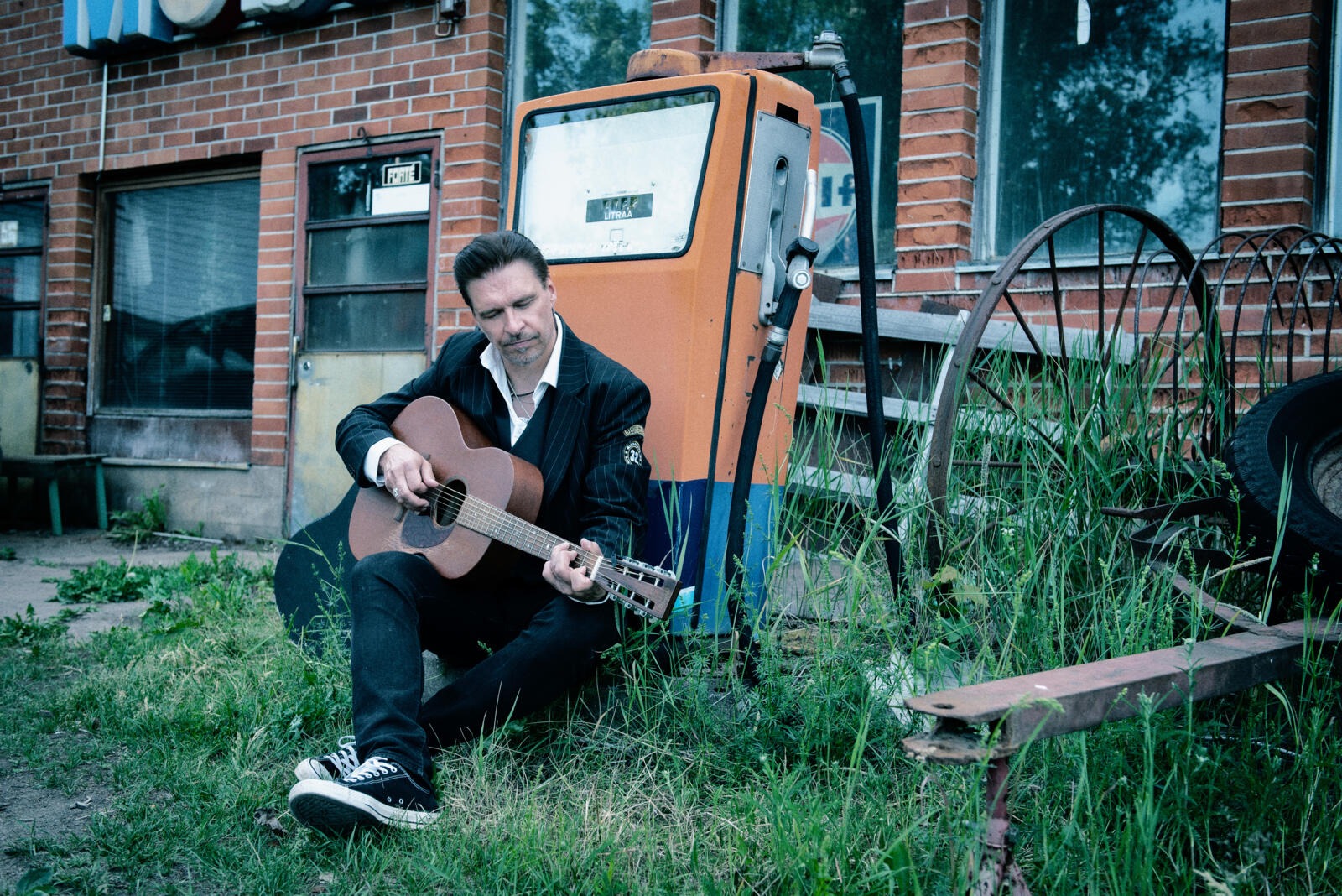 Man playing acoustic guitar while crouching outdoors on grass, brick building and vintage orange fuel pump in background.