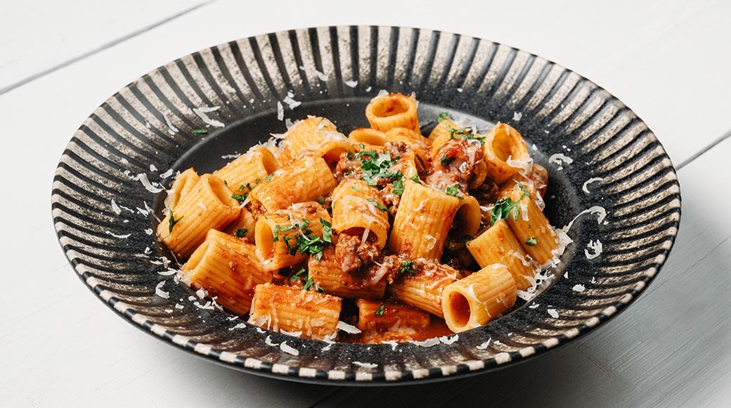 Rigatoni pasta lihakastmes, peal riivjuust ja hakitud petersell. Roog on mustal triibulisel taldrikul valgel laual.