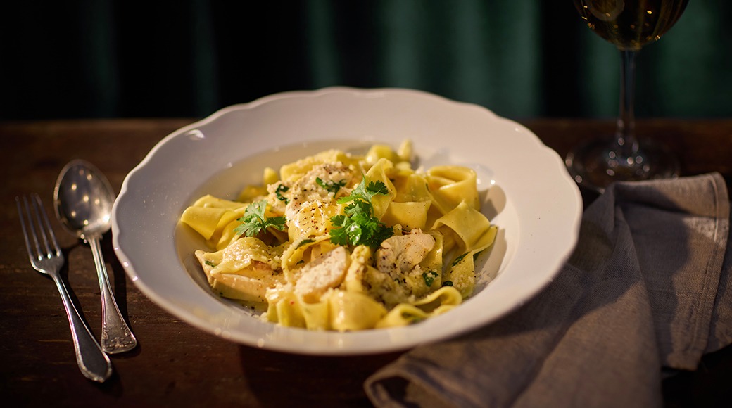 Valge kauss pasta ja kanaga, koriandriga kaunistatud, söögiriistad ja veiniklaas puidust laual.