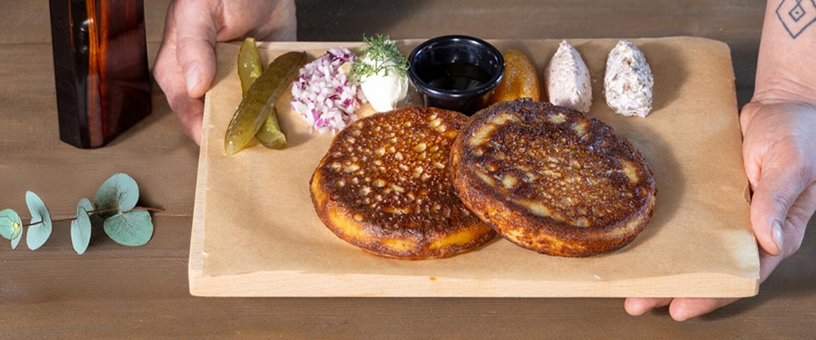 Bliniannos, jossa kaksi bliniä, suolakurkkuja, sipulisilppua, smetanaa ja kastiketta. Kädet pitelevät annosta.