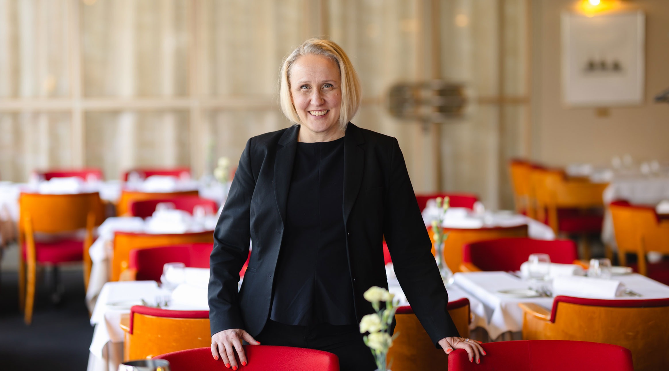 Nainen seisoo kahden punaisen pehmustetun tuolin välissä ravintolasalissa, jossa on pöydät katettu valkoisilla liinoilla. Tila on tasokas, lattia on tumma ja tasainen.