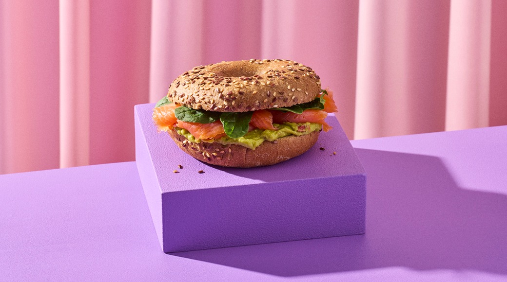 Siemenillä koristeltu bagel, jossa on lohta, avokadotahnaa ja pinaatinlehtiä. Bagel on violetin korokkeen päällä vaaleanpunaisen verhon edessä.