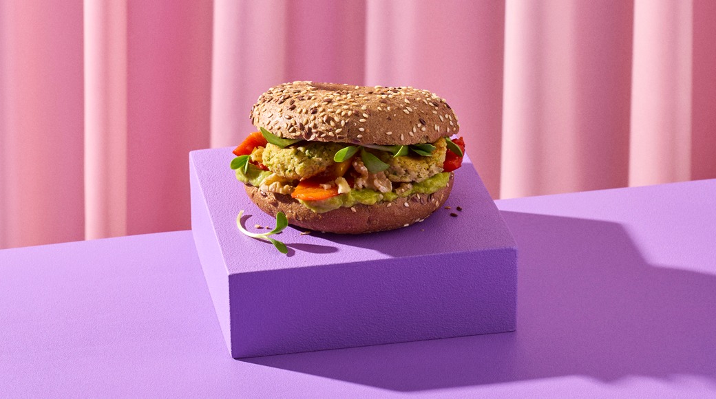 Seesaminsiemenillä koristeltu bagel, jonka välissä on avokadotahnaa, kasvispihvi, paahdettua paprikaa ja versoja. Bagel on violetin korokkeen päällä vaaleanpunaisen verhon edessä.