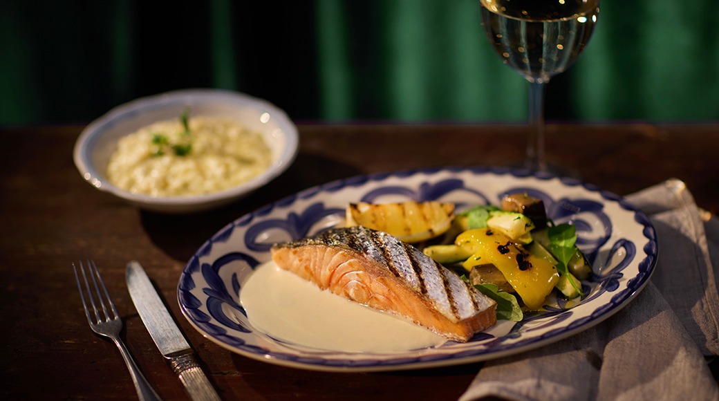 Grillad lax i ljus sås, grönsaker på en dekorerad blå-vit tallrik. En skål med ris och ett vinglas i bakgrunden.