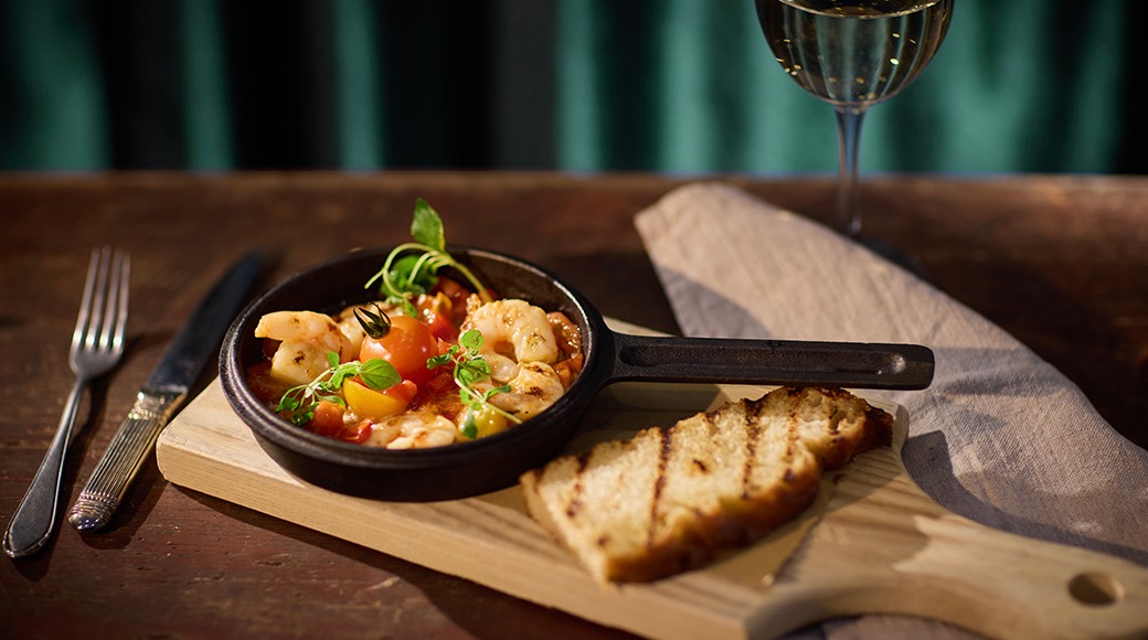 Malmpajas serveeritud krevettide ja tomatitega roog, puidusel alusel grillitud leiva ja veiniklaasiga.