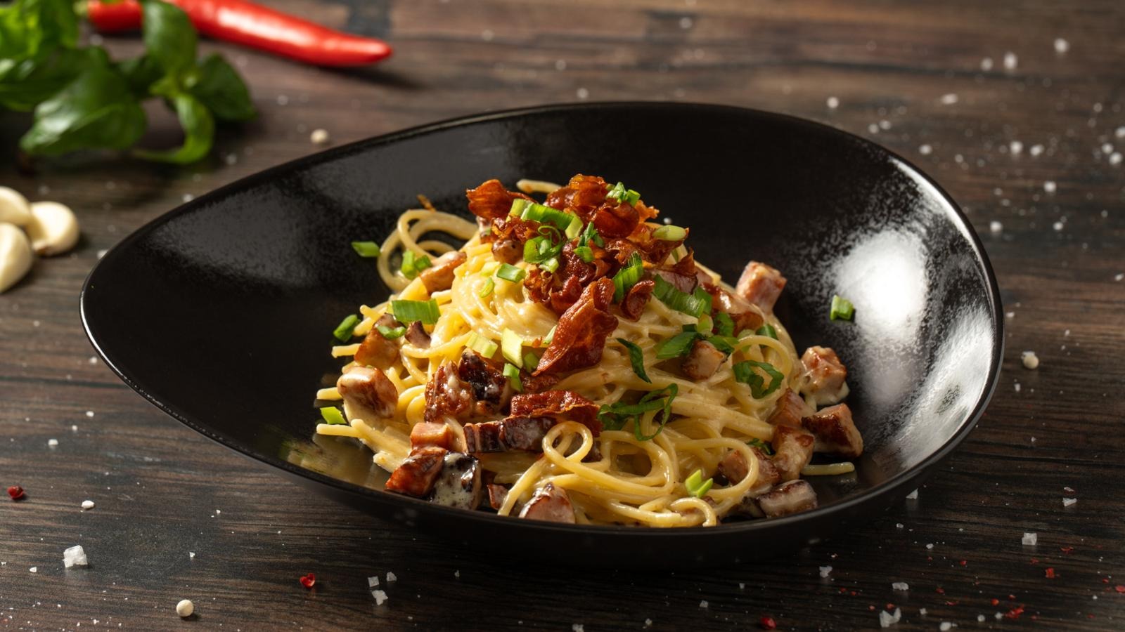 Mustassa kulhossa pastaa, jossa pekonia, sipulia ja lihaa. Ympärillä pöydällä chiliä, valkosipulinkynsiä ja mausteita.