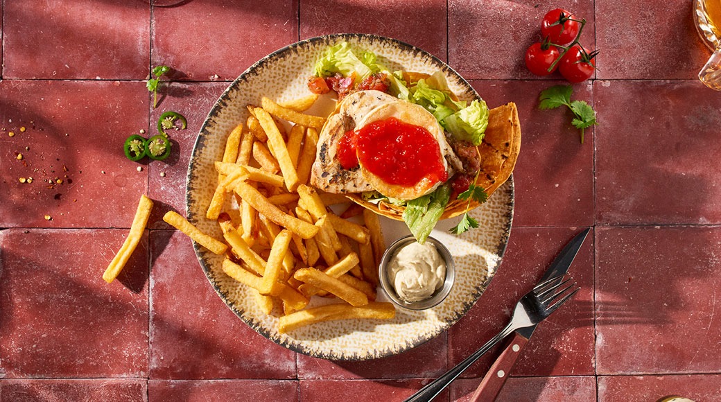 Taldrikul on friikartulid, tortillapõhine roog grillitud kana, salati ja punase salsaga ning väike kaste kausikeses. Kõrval on nuga ja kahvel punasel plaaditud laual.
