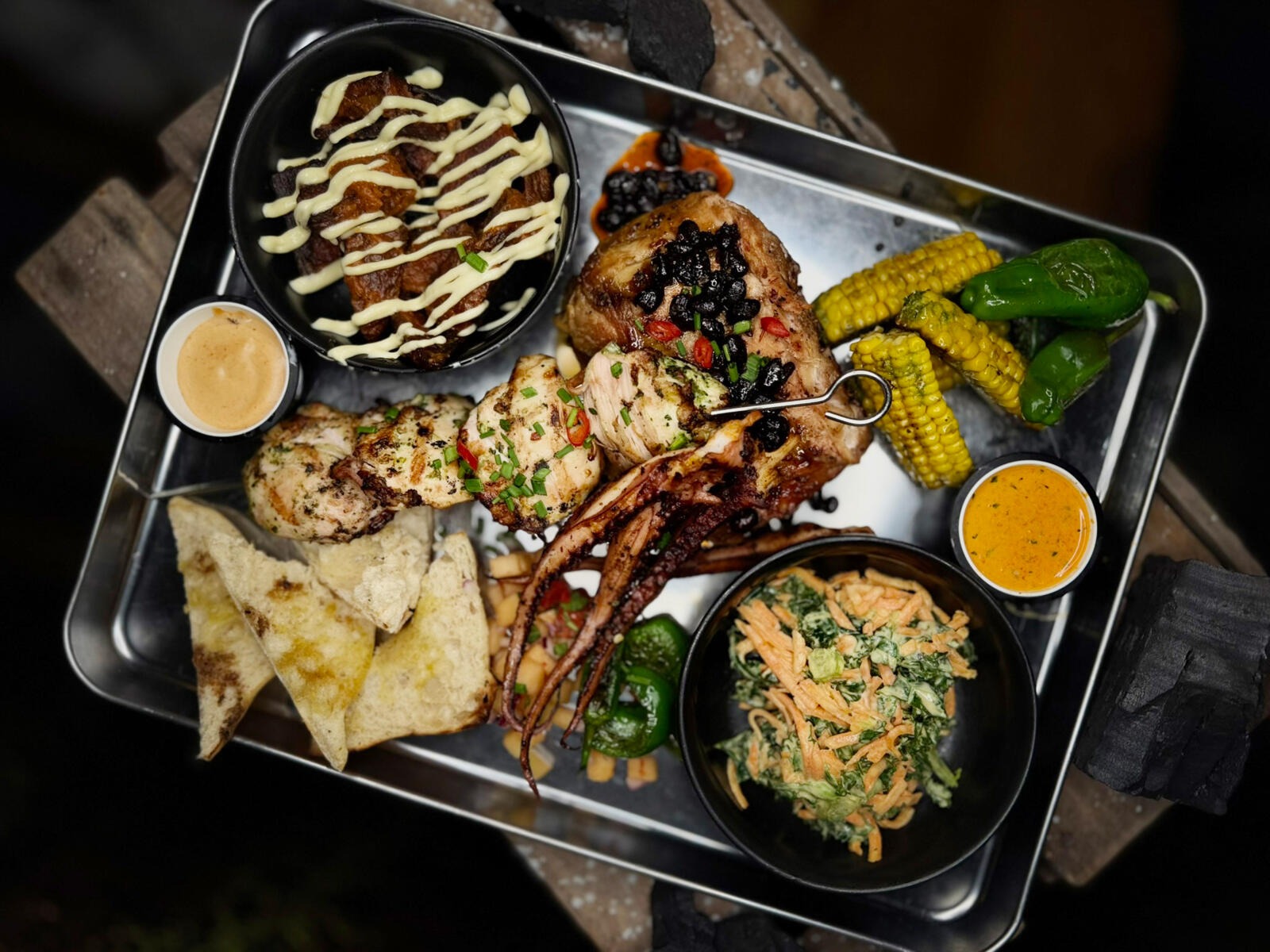 A meal served on a metal tray featuring chicken, grilled corn, peppers, tacos, salad and various sauces in small bowls.