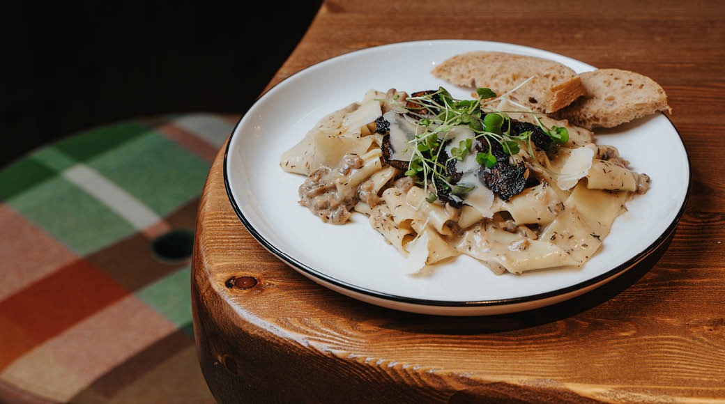 Lautasella pastaa sienikastikkeessa, koristeltuna versoilla ja tarjoiltuna leivän kanssa puisella pöydällä.