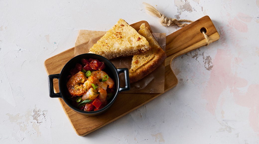 Small black cast iron dish of prawns in tomato sauce with spring onion. Garlic bread sits beside it on a wooden serving board.
