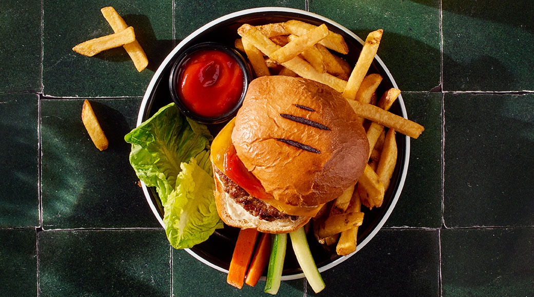 Burgeri, ranskalaisia, ketsuppia, salaattia sekä porkkana- ja kurkkutikkuja mustalla lautasella vihreällä laattapöydällä.