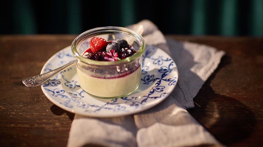 Väike klaaspurk pannacottaga, mille peal on värsked marjad, sini-valgel taldrikul tumedal puitlaual.