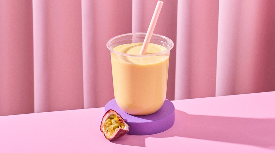 Keltainen smoothie kirkkaassa kupissa vaaleanpunaisen pillin kanssa violetilla alustalla. Vieressä puolikas passionhedelmä vaaleanpunaisella pinnalla.
