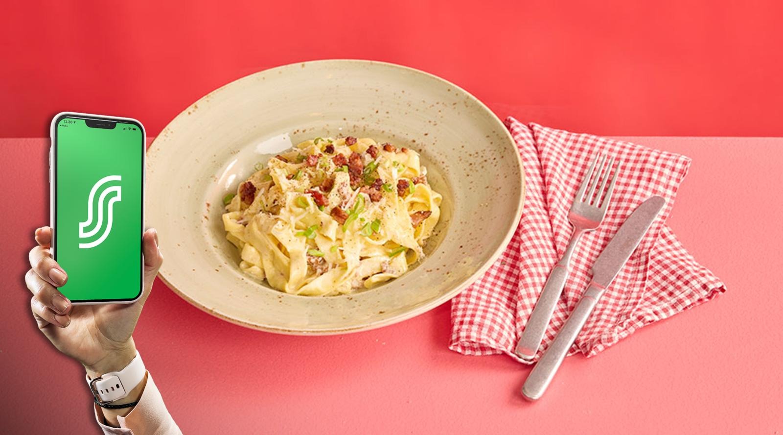 Käsi pitelee älypuhelinta, jonka vieressä on keraaminen lautanen pastaa, vihreää sipulia ja pekonia. Punavalkoinen ruudullinen lautasliina ja ruokailuvälineet vieressä.