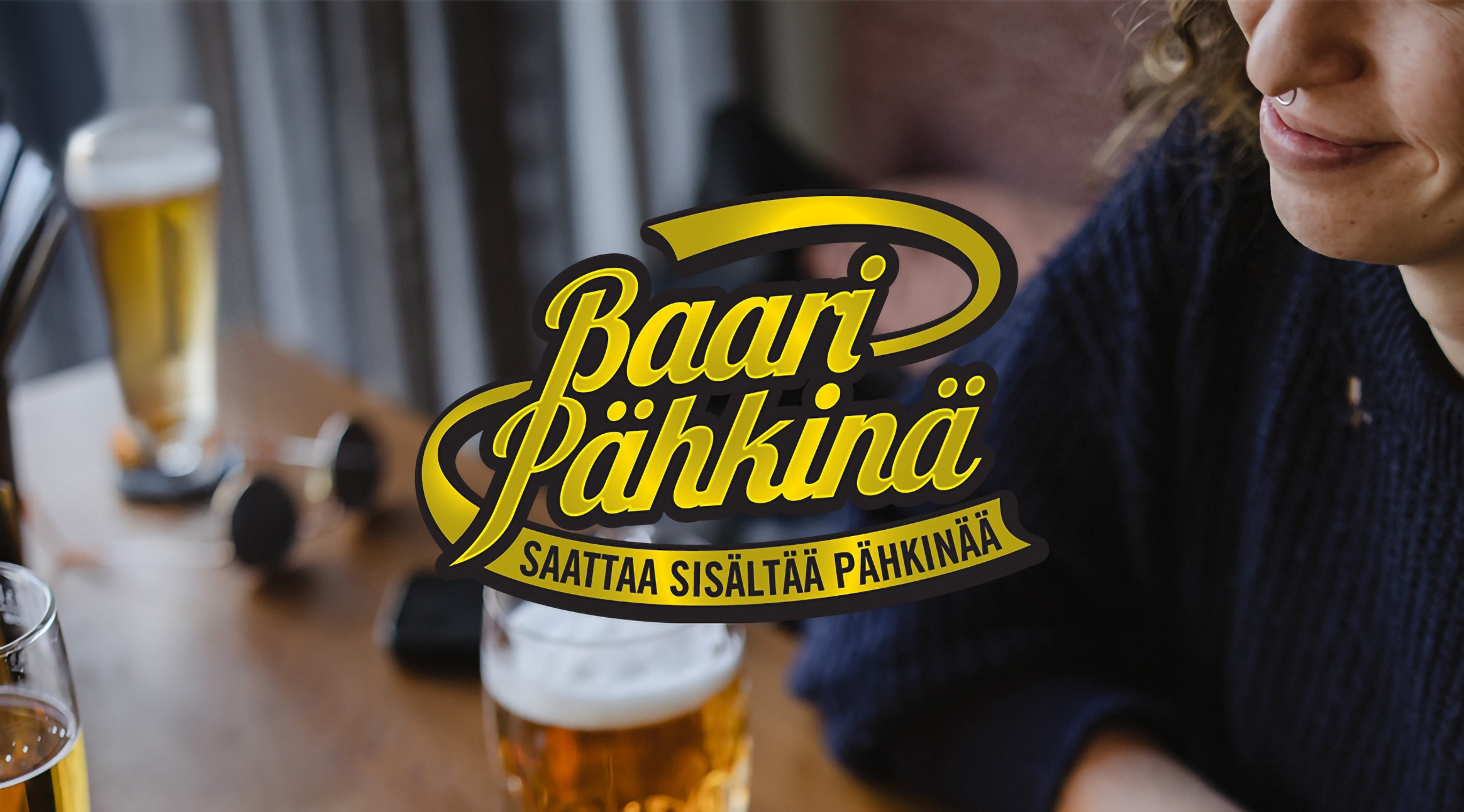 Nainen hymyilee ravintolapöydällä olevan oluttuopin ääressä. Kuvan keskellä on Baaripähkinä pubivisan logo.
