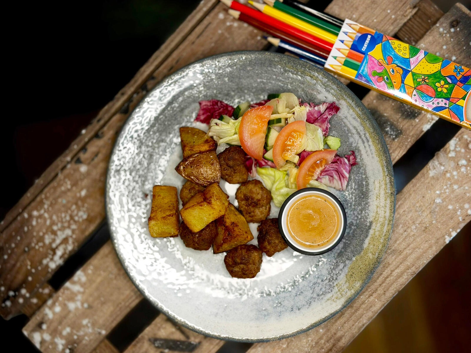 A meal on a ceramic plate: fried potatoes, meatballs, salad with tomatoes, and a dipping sauce. Colorful pencils sit nearby on the wooden table.