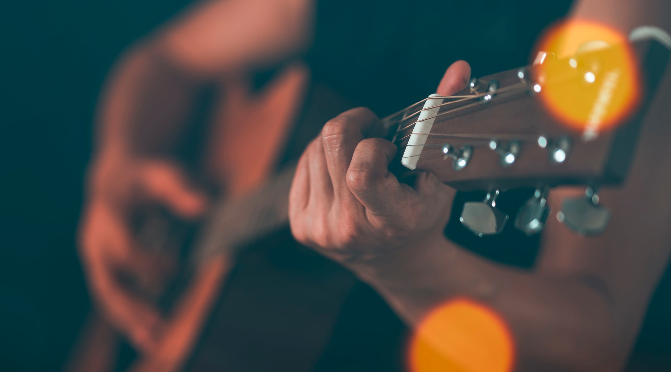 Hand spelar akustisk gitarr, fingrar på greppbrädan. Orange belysning syns i bakgrunden.