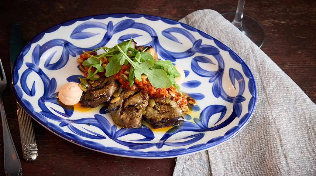 En portion stekt aubergine med tomatsås och rucola på en blå- och vitmönstrad tallrik, med rosa sås vid sidan.