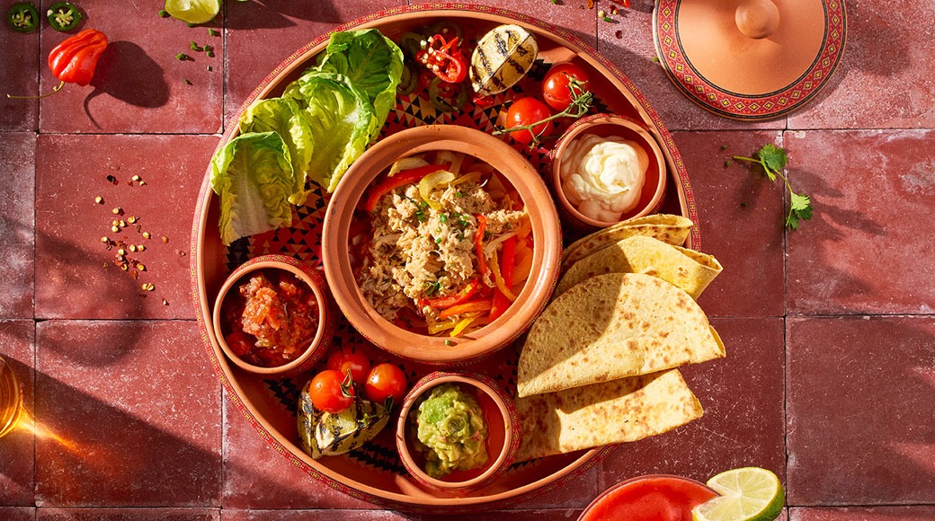 Terrakottalautasella tortillankolmioita, lihasekoitusta, salsaa, guacamolea, smetanaa, salaattia ja grillattuja vihanneksia punaisella laattalattialla.