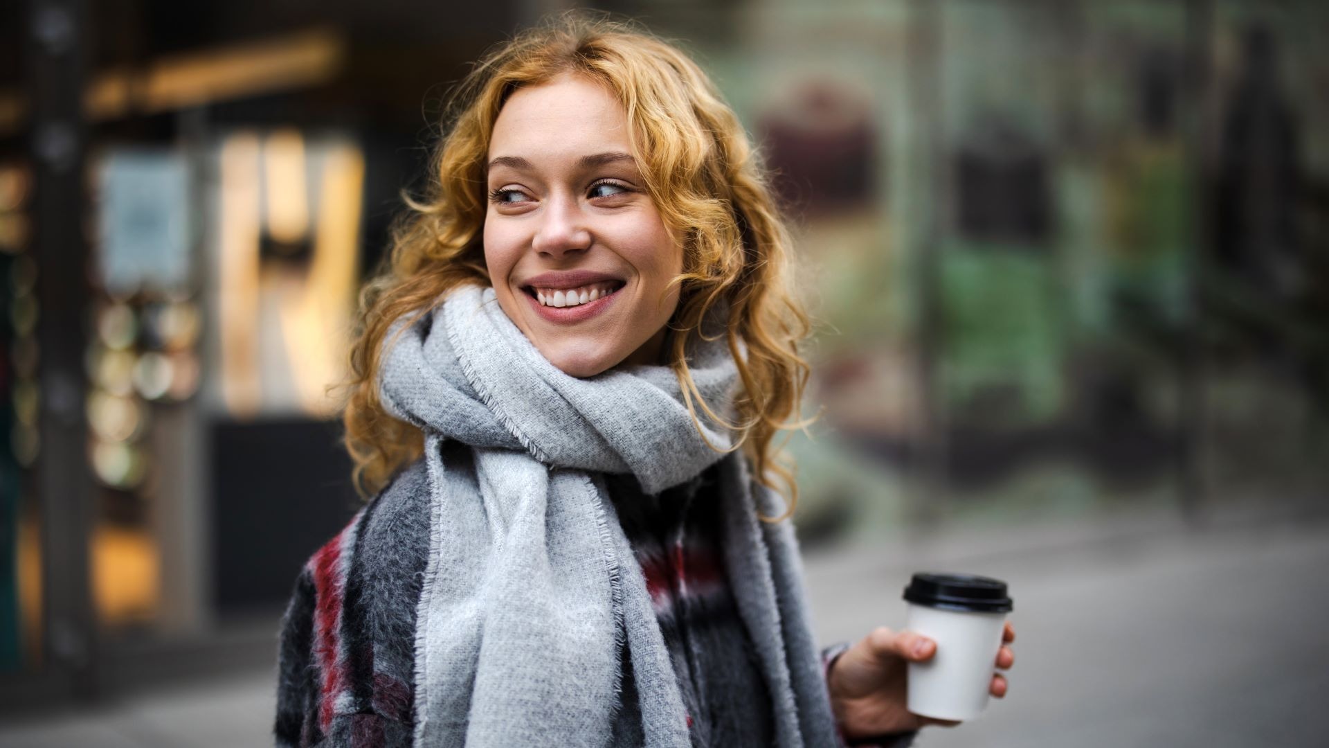 Person med lockigt hår ler utomhus, klädd i en grå halsduk och håller en take-away kaffekopp i handen.