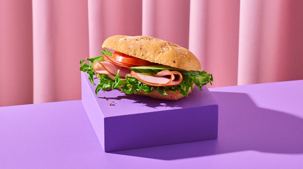 Kinkku-salaattisämpylä, jossa on tomaattia ja kurkkua, purppuranpunaisella korokkeella vaaleanpunaista verhoa vasten.