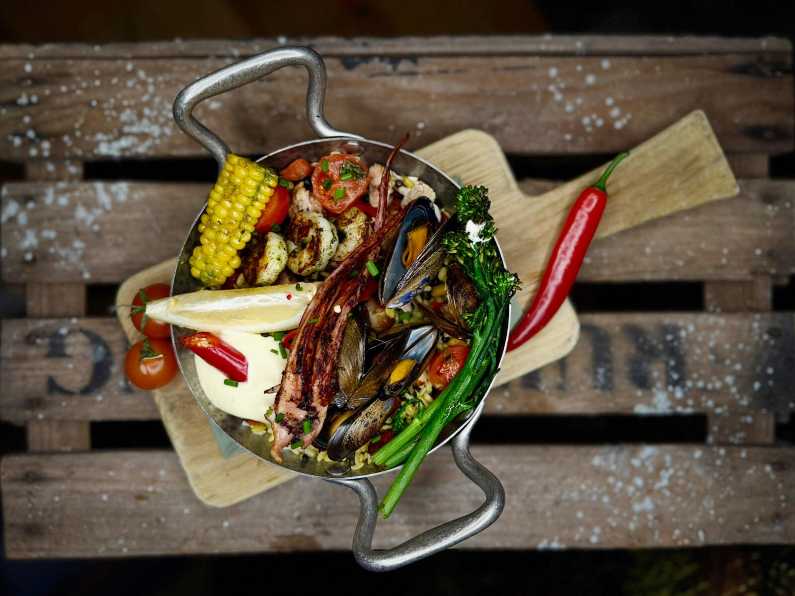 Seafood paella in metal pan with mussels, shellfish, corn and vegetables on wooden board.
