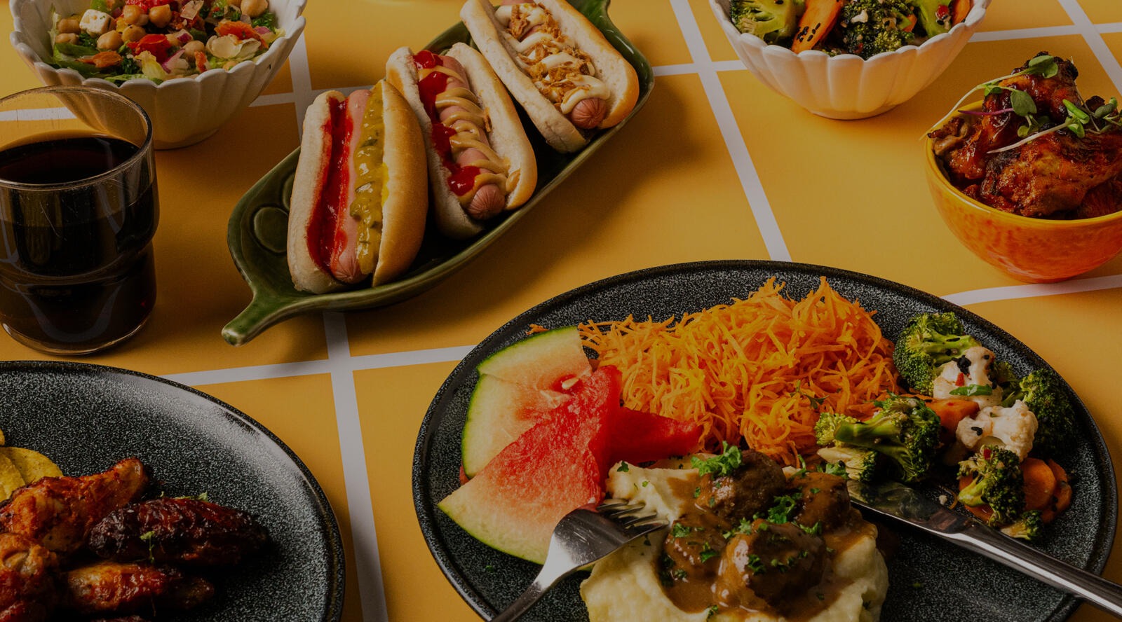 Ett bord med olika maträtter: varmkorvar, sallad, riven morot, vattenmelon, köttbullar med potatismos och kycklingbitar. Drycker i bakgrunden.