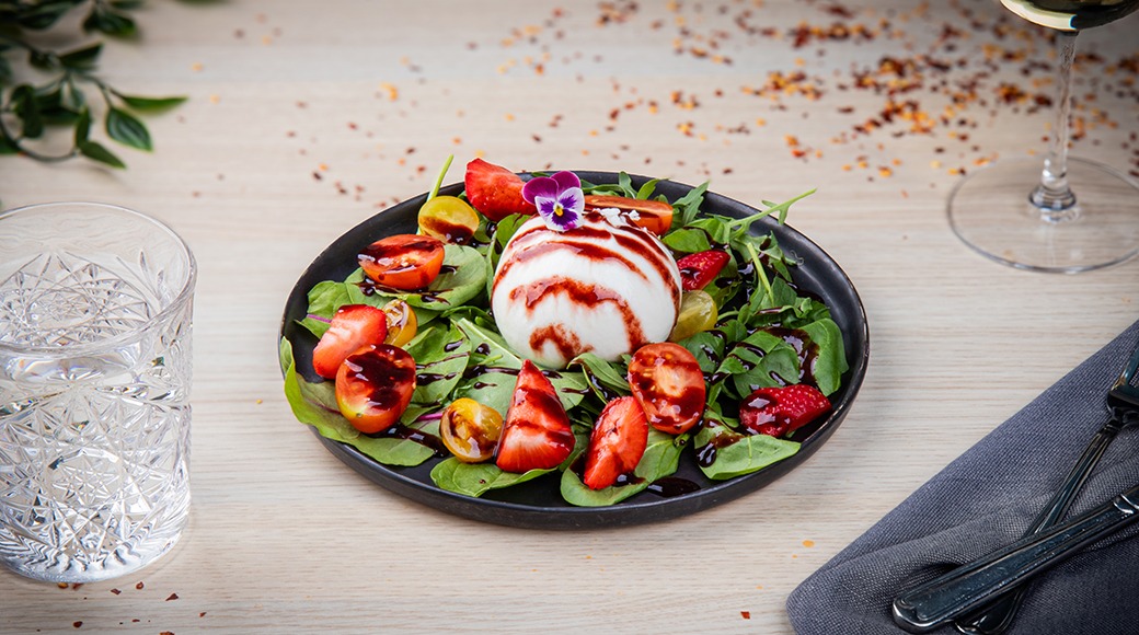 Salaattilautanen, jossa on burrata-juustoa, mansikoita, tomaatteja ja vihreitä lehtiä, aseteltu vaalealle pöydälle.