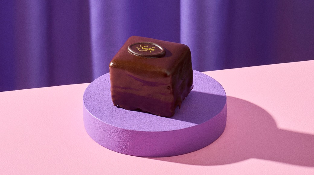 Kiiltävä suklaakakkupala, jonka päällä on suklaakoriste, violetilla korokkeella vaaleanpunaisella pöydällä. Taustalla on violetti verho.