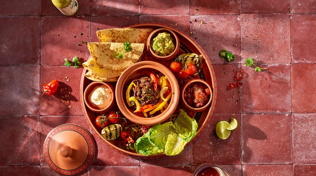 Fajitasöök terrakotasalvril: liha ja paprika, tortillad, salat, guacamole, salsa ja hapukoor. Kõrval on laim ja tšillid.