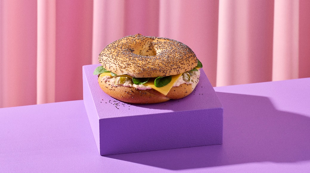 Poppy seed bagel filled with cream cheese, a cheese slice and spinach leaves. It sits on a purple block in front of a pink curtain.