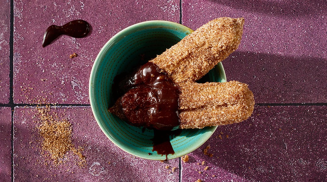 Kaksi sokeroitua churroa dipattuna suklaakastikkeeseen turkoosissa kulhossa violetilla laattapinnalla.