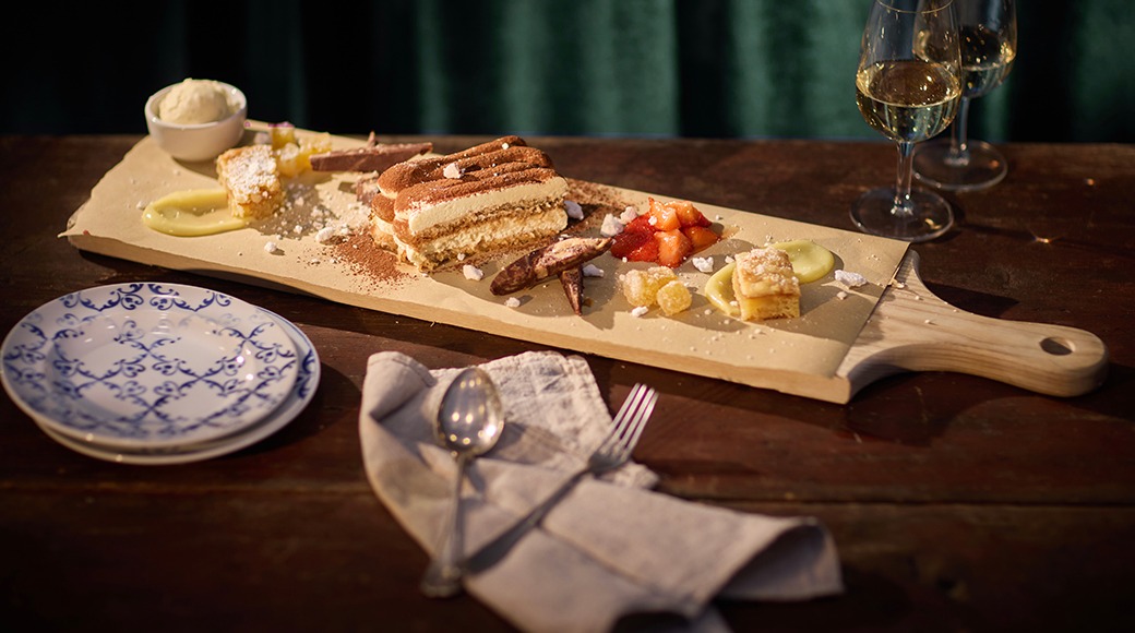 Träbricka med efterrätter, tiramisu i mitten, vitvinsglas i bakgrunden och en blåvit tallrik på bordet.