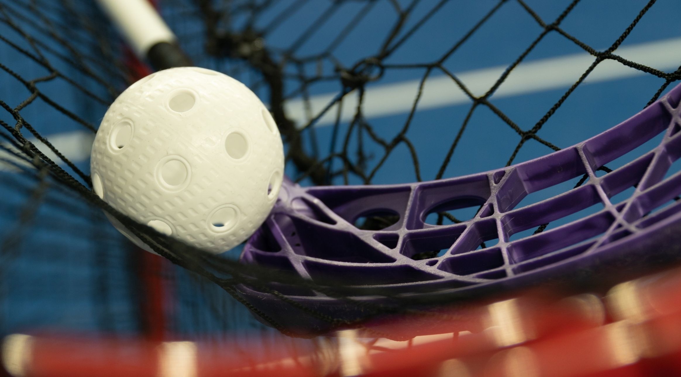 White floorball and purple stick blade against a black goal net. A blue indoor court is blurred in the background.