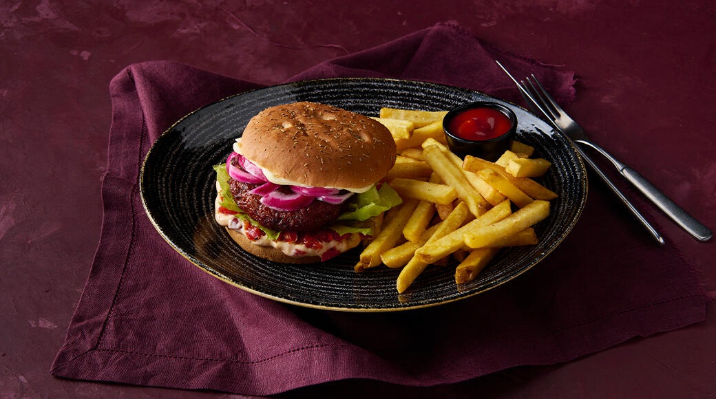 Burger, French fries and a small bowl of ketchup on a black plate. The plate sits on a burgundy cloth napkin, with a fork and knife beside it.