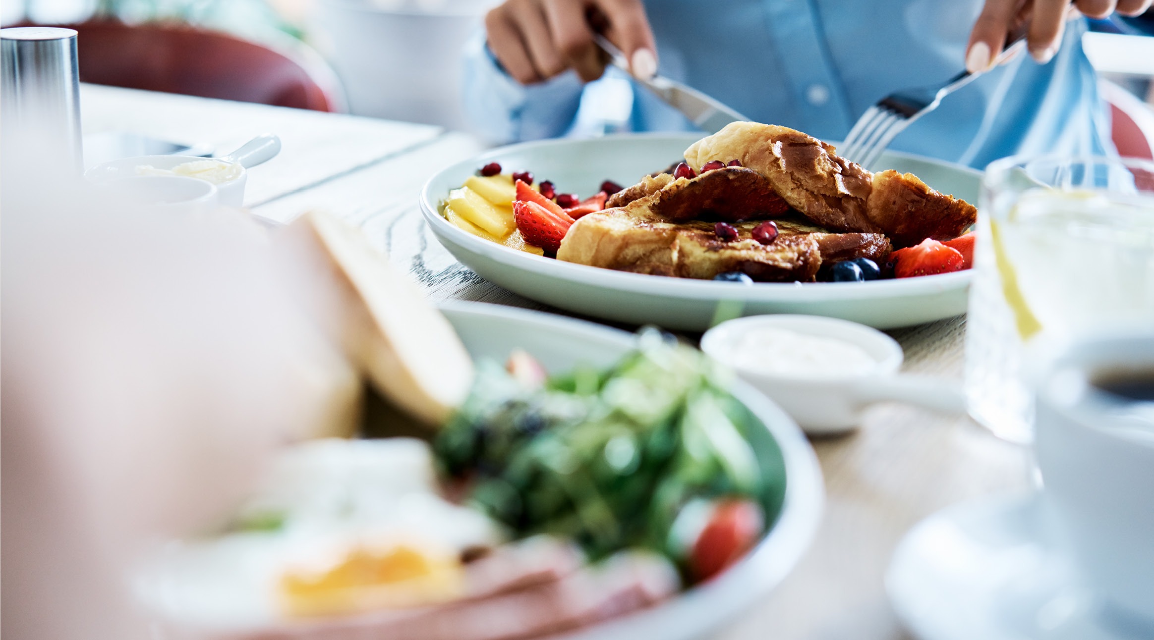 Person cutting French toast with fork and knife, garnished with berries and fruit.