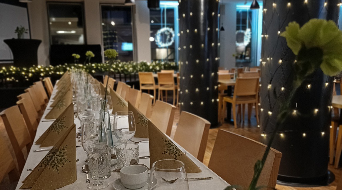Pitkä katettu juhlapöytä, jossa on lasit, lautasliinat ja vihreä neilikka. Pylväitä koristavat valonauhat. Lattia on puuta.