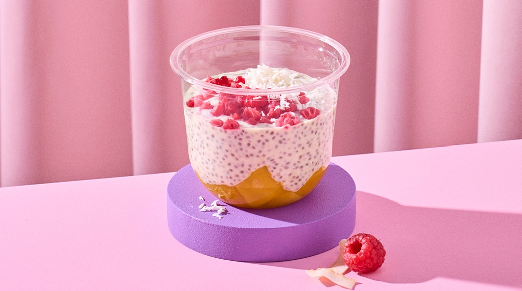 Kirkas kulho, jossa chiavanukasta, mangokerros sekä vadelmamurskeita ja kookoshiutaleita päällä. Kulho on violetilla alustalla vaaleanpunaista taustaa vasten, vieressä vadelma.