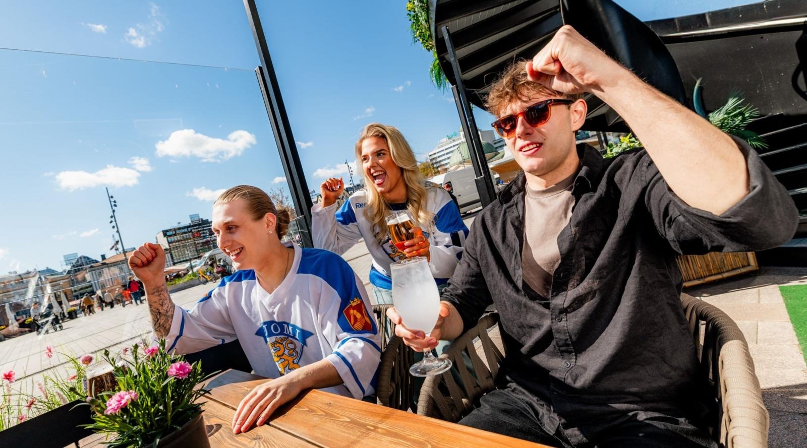 Kuvassa kaksi henkilöä Suomi-kisapaidoissa ja yksi muu fani seuraamassa ulkona BAR W:n terassilla urheilua screeniltä.