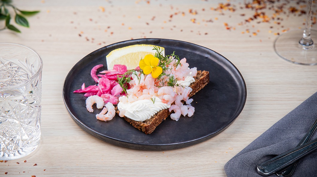 Katettua pöytää ravintolassa. Lautasella rye-leipää, koristeellisesti aseteltu katkarapu, vaaleanpunainen sipuli ja kukkia.